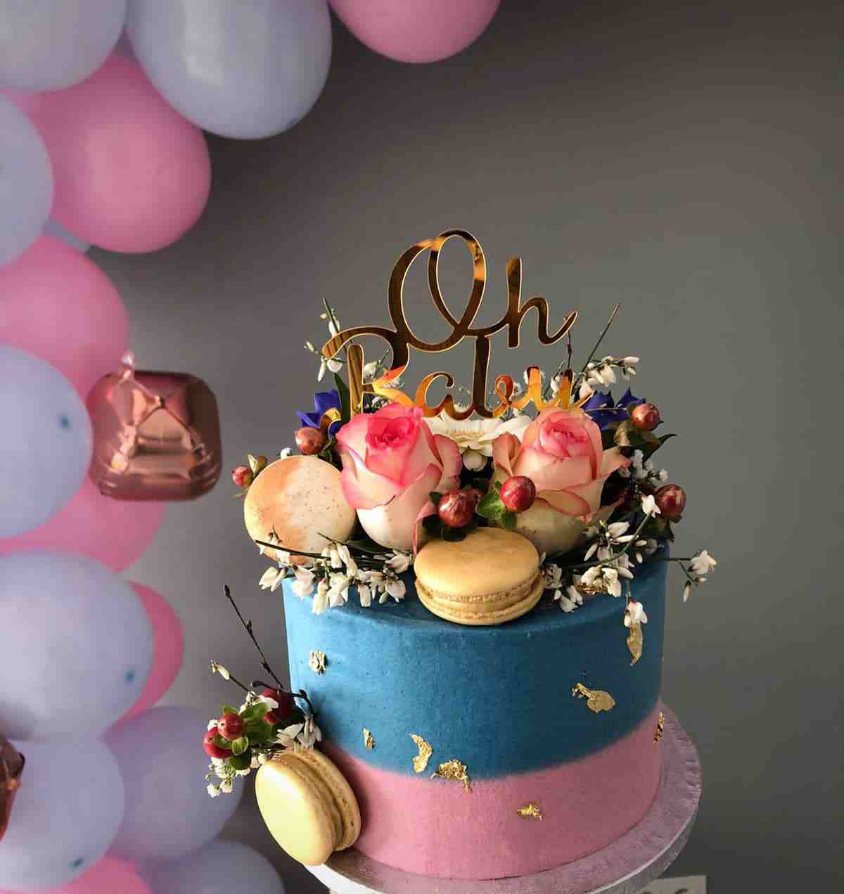 Gender reveal taart in blauw en roze met gouden Oh Baby-topper, rozen en macarons bij een ballonnenboog