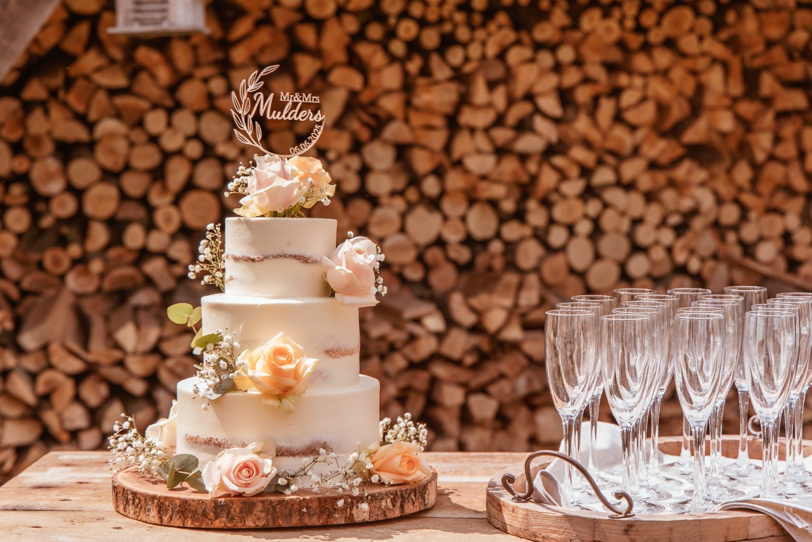 Semi-naked bruidstaart met perzikkleurige rozen en gipskruid voor een houtstapel, naast champagneglazen