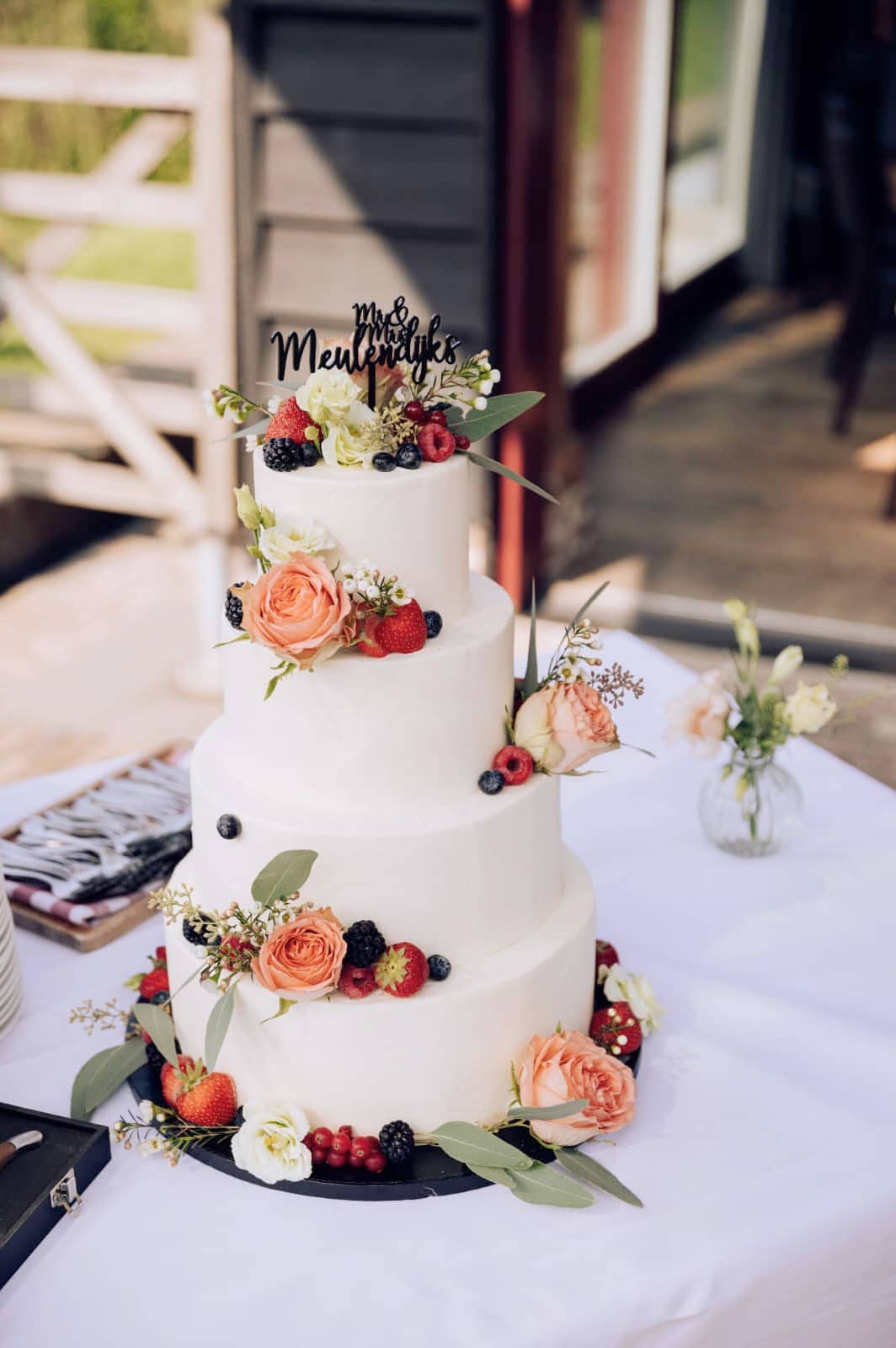 Vierlaagse witte bruidstaart met vers fruit, zalmroze rozen en Mr & Mrs-topper op een feesttafel